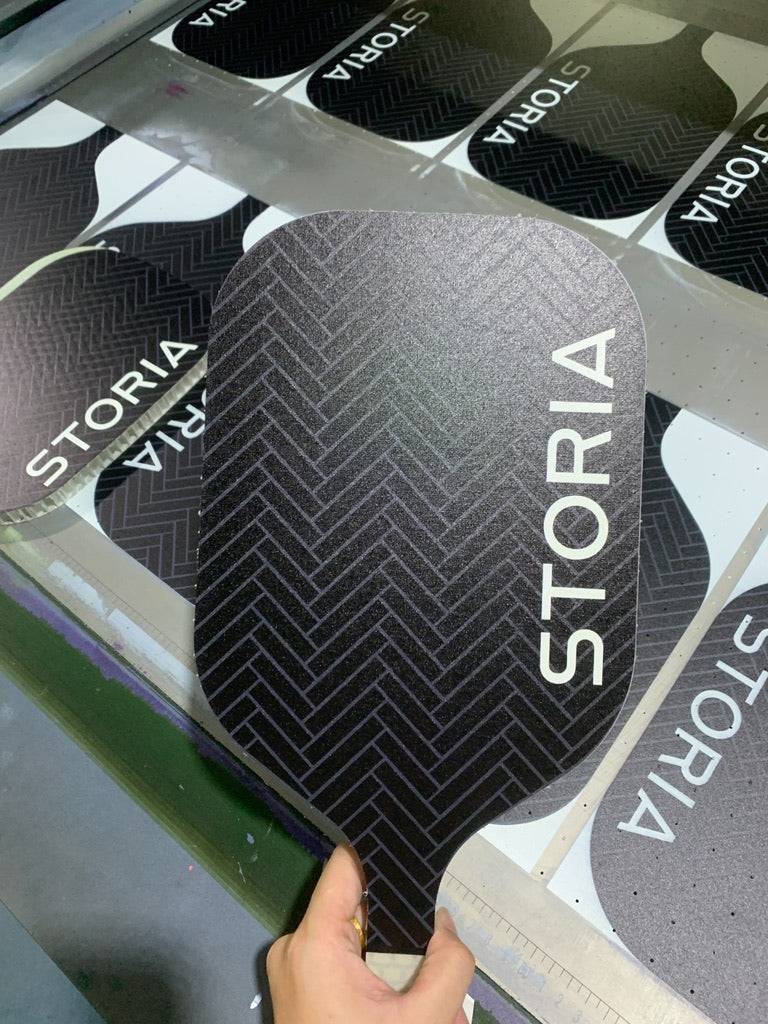 Custom black paddle with 'STORIA' branding held by a hand against a display of similar paddles.