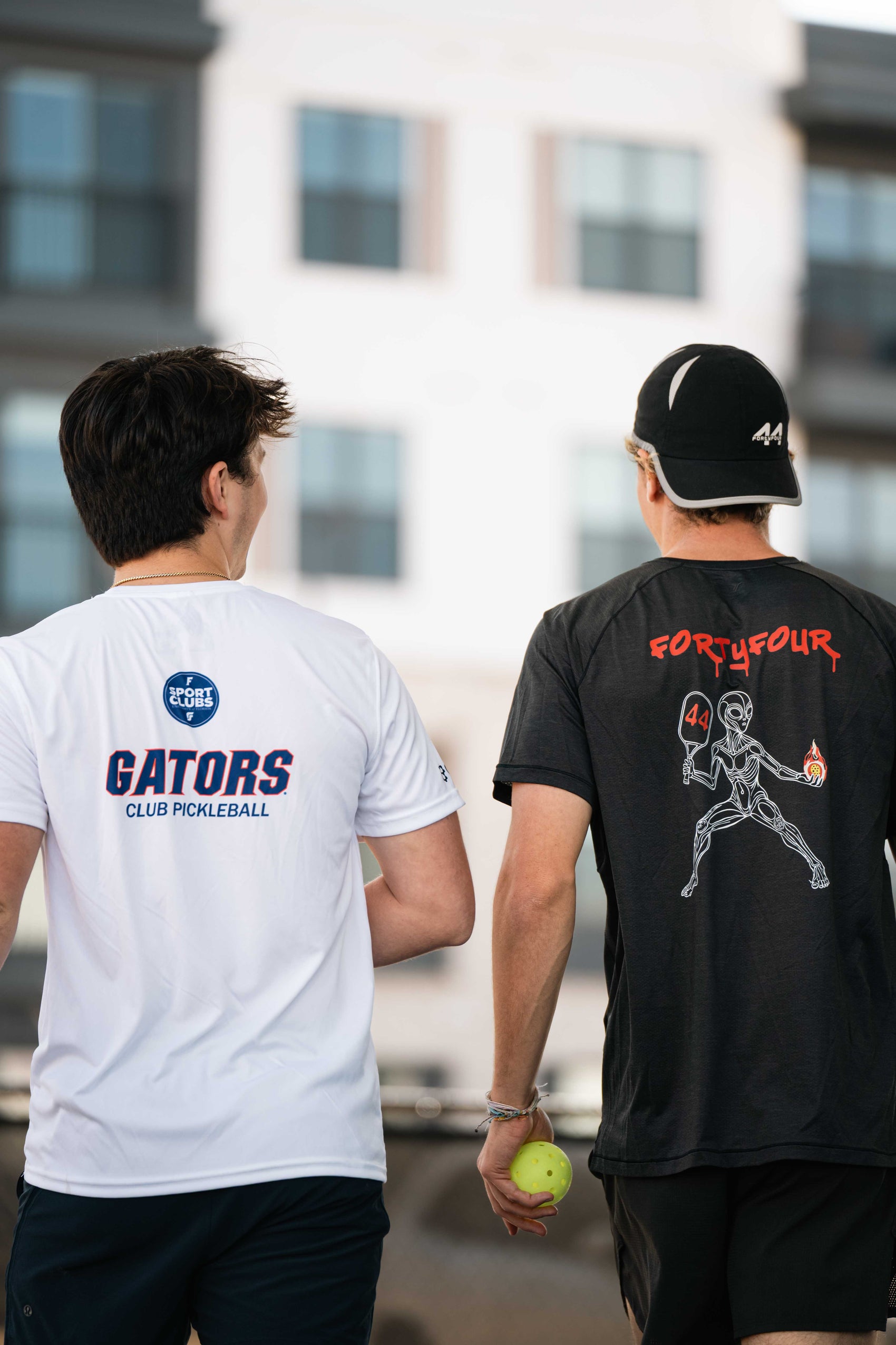 Two people playing pickleball, one wearing custom shirts: a white 'Gators Club Pickleball' shirt and the other in a black 'Four Four' shirt.
