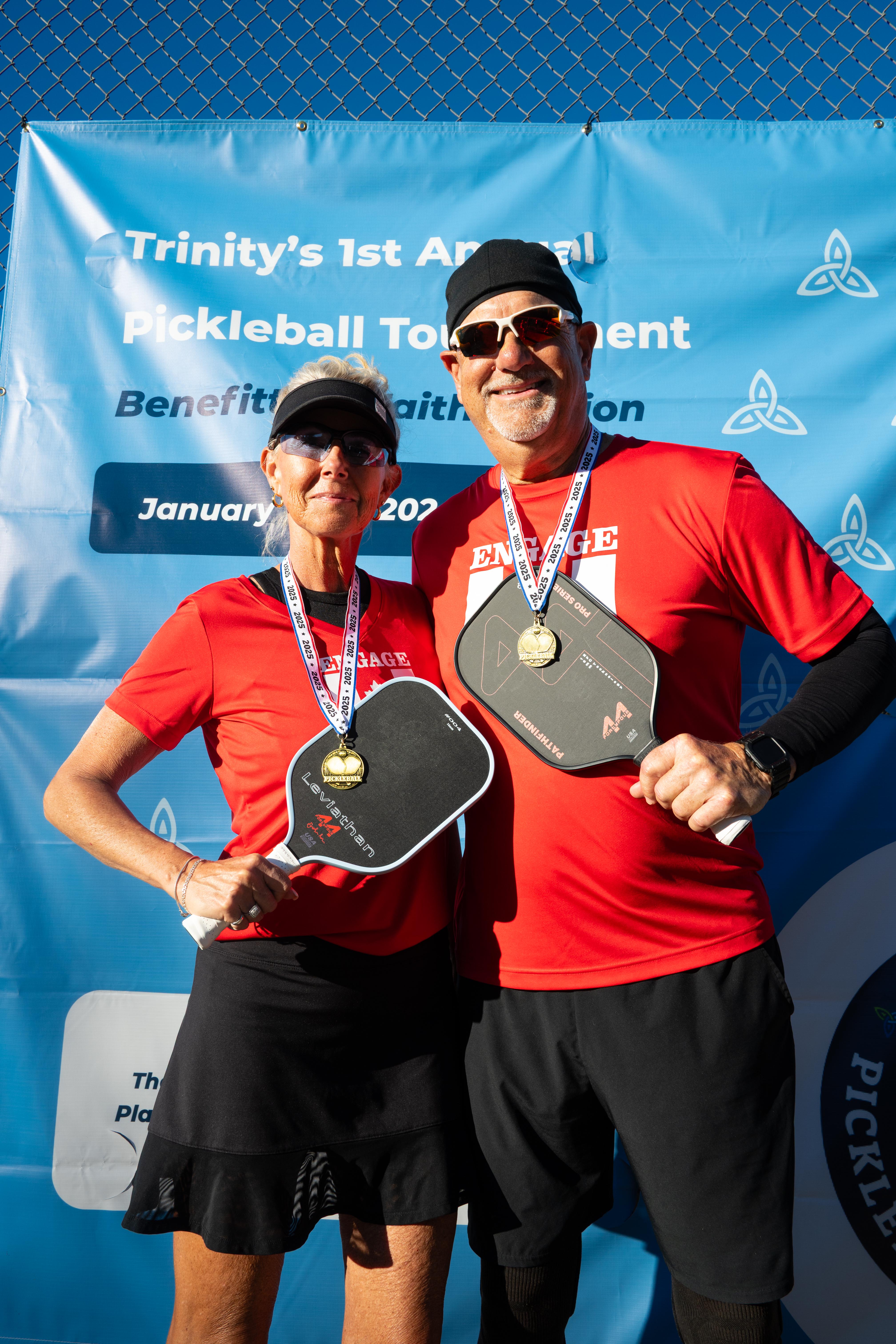 Two people holding pickleball paddles in front of a blue banner with text